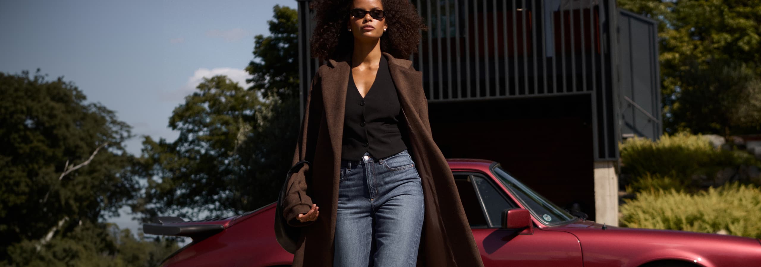 Model is wearing a brown coat over a black vest and blue jeans.