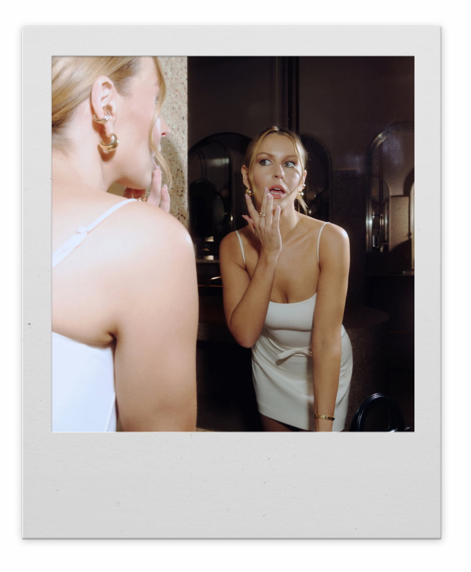 A blonde woman in a white mini dress checks her make up in a mirror.