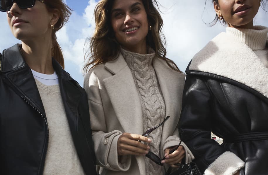 Models in white and grey winter coats sit at a table in a cafe.