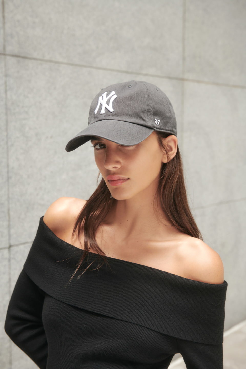 A model wears a grey New York cap with a black off-the-shoulder shirt.