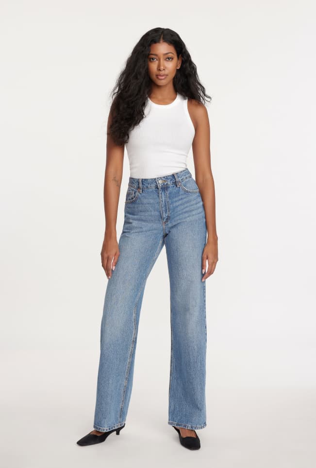 Model is wearing a white tank top and blue wide leg jeans.