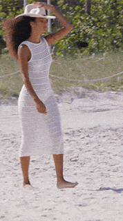 A woman with dark curly hair in a white open knit maxi dress lifts a white cowboy hat from her obscured face.