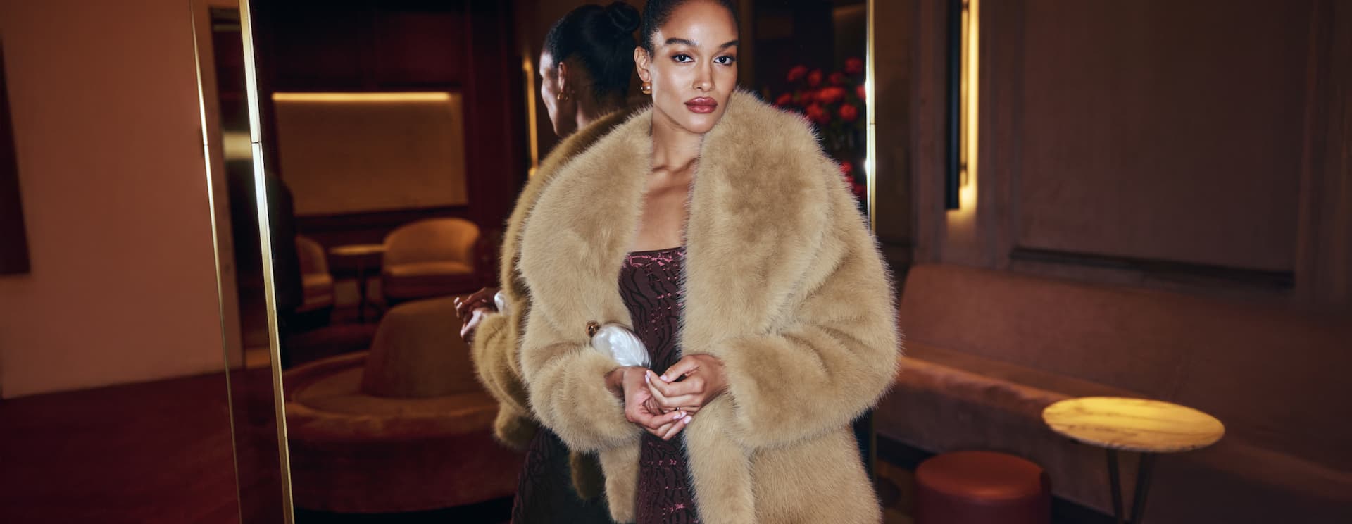 Model is wearing a beige faux fur jacket over a burgundy maxi dress with a velvet floral print.