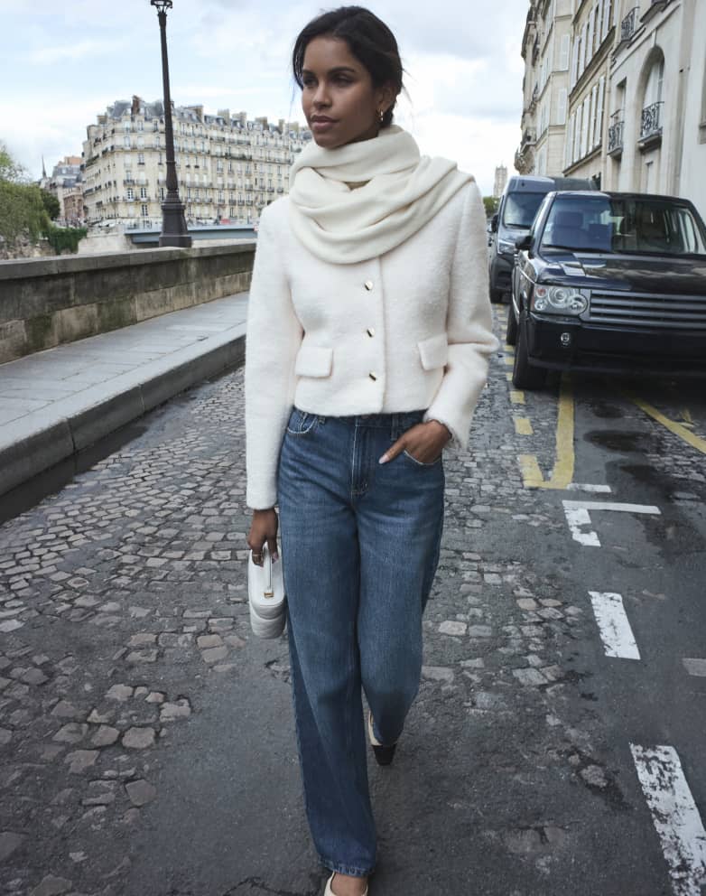 Model is walking down a cobblestone street wearing a white scarf and cardigan, and blue jeans.