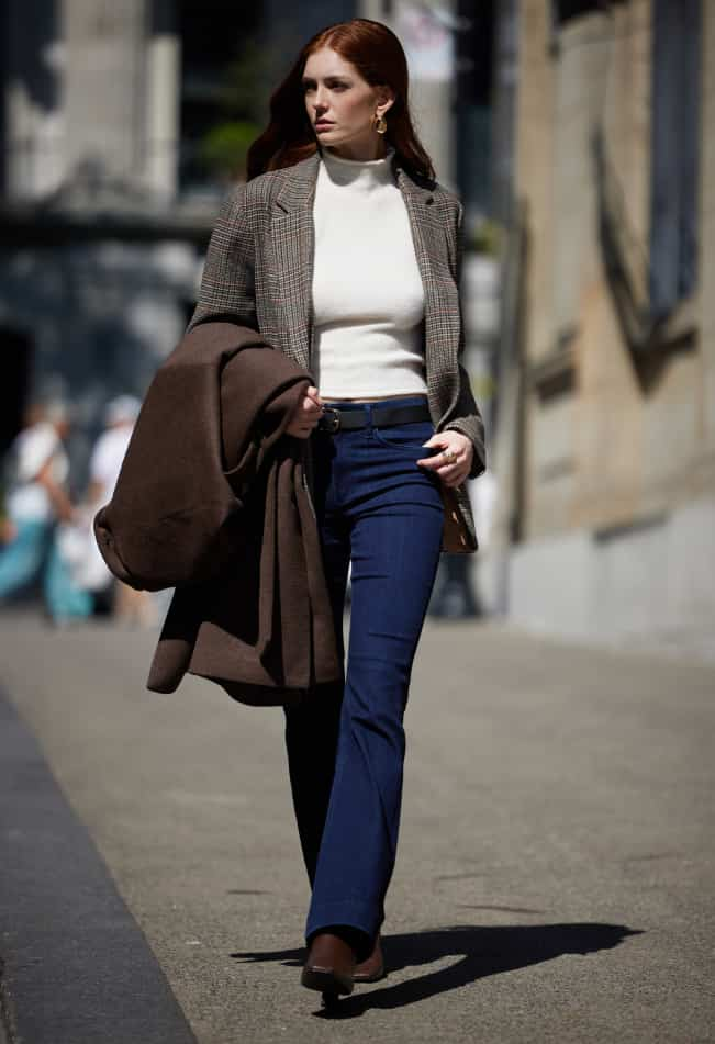 Model is wearing a brown tweed blazer over a white mock neck top and indigo bootcut jeans.