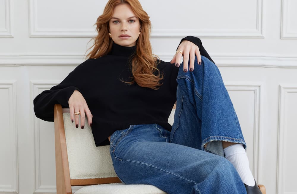 Model reclines in a black turtleneck and blue jeans.