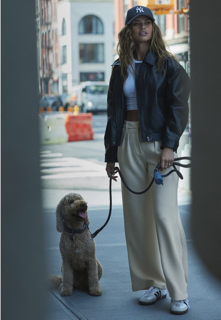 Model is wearing a navy cap and a black faux leather jacket over a white T-shirt and beige pants.